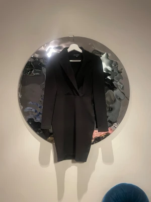 Svart kavajklänning från amisu  - Stilren svart kavajklänning från amisu med lång ärm och v-ringad omlottfront. Klänningen har en figurnära passform och klassisk krage, perfekt för en trendig look. Materialet känns mjukt och följsamt.