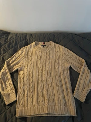 Beige kabelstickad tröja Tommy Hilfiger - Kabelstickad beige tröja från Tommy Hilfiger med rund halsringning och diskret logga på bröstet. Tröjan har långa ärmar, ribbade muddar och nederkant. Perfekt för dig som gillar klassisk och stilren design. Tröjan är storlek 164, så ungefär xs 