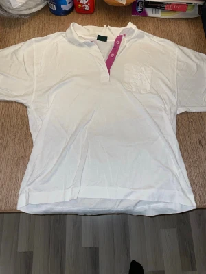 Vit piké med rosa knappslå från Lyle & Scott - Säljer en vit pikétröja med kort ärm och klassisk krage. Tröjan har en lila knappslå med tre knappar och en bröstficka på vänster sida med loggan Lyle & Scott på. Perfekt för en avslappnad och stilren look. Materialet känns mjukt och bekvämt.