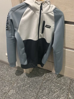 Grå och vit hoodie från McKenzie - Snygg hoodie från McKenzie i grått och vitt med dragkedja framtill, två sidofickor med dragkedja och en bröstficka. Stor logga på ärmen och patch på bröstet. Tillverkad i mjukt material, perfekt för chill eller streetwear.
