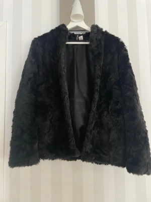 Svart fuskpälsjacka - Snygg svart fuskpälsjacka med mjuk och fluffig känsla. Jackan har rak passform, långa ärmar och är helsvart både på utsidan och fodret. Perfekt för att hålla värmen under kalla dagar och ger en trendig look.