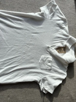 Vit t-shirt i frotté från OAS - Säljer en vit t-shirt från OAS i mjuk legendarisk frotté. T-shirten har klassisk krage, korta är sopade är sopade ärmar och en liten bröstficka. Perfekt för chill dagar och enkel att sopade till shorts eller jeans.