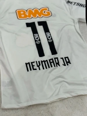 Vit Santos Neymar Jr matchtröja - Säljer en vit Santos FC matchtröja från Nike med Neymar Jr och nummer 11 på ryggen sopm har svarta tryck. Tröjan har flera sponsorloggor, bland annat BMG i orange och svart, samt klubbmärke på bröstet. Klassisk rund hals och korta ärmar, perfekt för fotbollsfans.