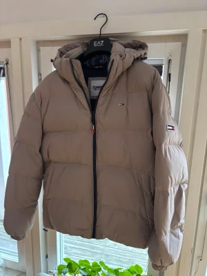 Beige pufferjacka från Tommy Hilfiger - Säljer en beige pufferjacka från Tommy Hilfiger med huva och dragkedja framtill. Jackan har logotypdetaljer på bröstet. Knappt använd!