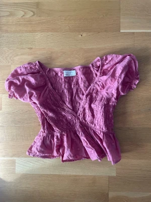 Rosa omlottopp från Urban Outfitters - Supersöt rosa omlottopp från Urban Outfitters med puffärmar. Toppen har volang nedtill och en croppad passform som ger en trendig look. Köpt 2022 och sparsamt använd. 
