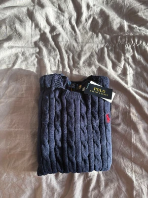 Mörkblå kabelstickad tröja Polo Ralph Lauren - Mörkblå kabelstickad tröja från Polo Ralph Lauren med rund halsringning och röd broderad logga på bröstet. Tröjan har långa ärmar och ribbade muddar vid ärmslut och nederkant. Klassisk och tidlös design i mjukt material. Mått:  Längd: 60 cm Axelbredd: 35 cm