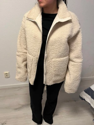 Krämvit teddyjacka med dragkedja - Mysig krämvit teddyjacka med hög krage och dragkedja framtill. Jackan har två stora fickor på sidorna och är tillverkad i fluffigt teddy-material som ger en avslappnad och trendig look. Perfekt för kalla dagar när du vill vara både varm och snygg.