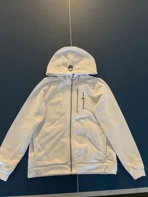 Vit hoodie från Sail Racing - Säljer en vit hoodie från Sail Racing med dragkedja framtill och en bröstficka med logga. Hoodien har huva och är tillverkad i ett mjukt material med fleece på insidan. Perfekt för dig som gillar stilren och sportig look.