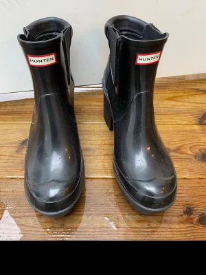 Svarta gummistövlar med klack från Hunter - Säljer ett par svarta gummistövlar från Hunter med bred klack och elastiska paneler på sidorna. Stövlarna har en klassisk logga framtill och är tillverkade i slitstarkt gummi. Perfekta för regniga dagar och ger en cool look till din outfit.
