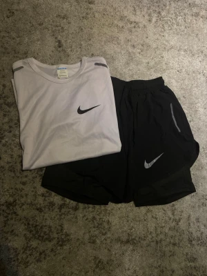 Nike träningsset t-shirt och shorts - Säljer ett sportigt träningsset från Nike med en ljusgrå t-shirt och svarta shorts. Båda plaggen har den klassiska Nike-loggan i vitt. T-shirten har rund hals och korta ärmar, shortsen har resår i midjan och reflekterande detaljer. Perfekt för gym eller löpning. Strl S, passar dig runt 160 lång.