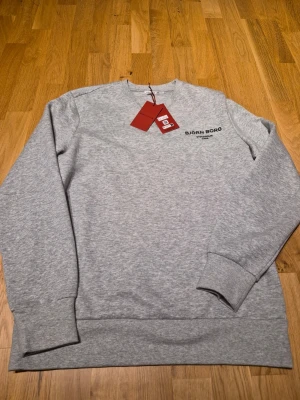 Grå sweatshirt från Björn Borg - Snygg grå sweatshirt från Björn Borg med svart tryck på bröstet där det står 'BJÖRN BORG STOCKHOLM 1956'. Tröjan har rund hals, långa ärmar och ribbade muddar. Perfekt för en avslappnad och stilren look. Storlek Large men är mer som Medium.  Tröjan är helt Ny. Då den är för liten för mig.