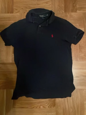 Svart pikétröja från Polo Ralph Lauren - Klassisk svart pikétröja från Polo Ralph Lauren med röd broderad logga på bröstet. Tröjan har korta ärmar, krage och knappar vid halsen. Perfekt för en stilren och avslappnad look.