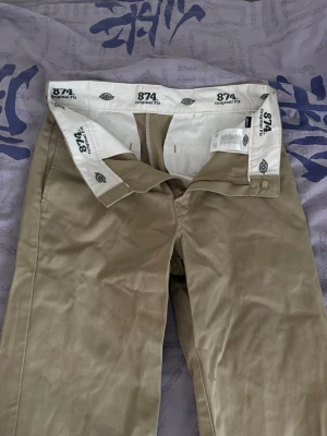 Beige Dickies 874 chinos - W31L32 Klassiska beige Dickies 874 chinos med rak passform och hög midja. Byxorna har en stilren look med diskreta fickor och slitstarkt material, perfekt för en clean och avslappnad stil. Materialet är en bomulls- och polyesterblandning.