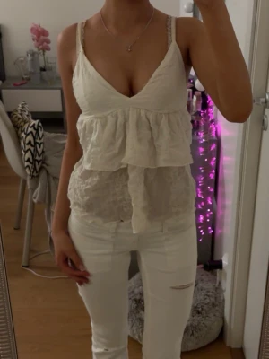 Vit volangtop från Gina Tricot - Supersöt vit linnetopp från Gina Tricot med volangdetaljer och djup v-ringning. Toppen är ärmlös med tunna axelband och har en luftig, lätt struktur i materialet.