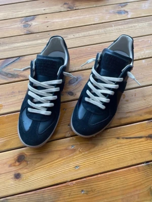 Svarta Maison margiela gats - Säljer ett par svarta sneakers från Maison Margiela med beige gummisula och vita snören. Skorna har en klassisk låg profil och är tillverkade i en mix av läder och mocka. Perfekt för dig som vill ha en stilren och tidlös look.