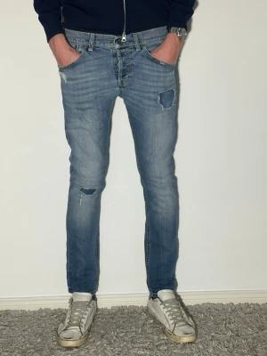 Blå dondup jeans med schyssta slitningar skinny fit - Säljer ett par blå jeans med slim fit och slitningar på både lår och knä. Jeansen har patchdetaljer och en klassisk femficksmodell. Perfekta för en avslappnad och trendig look.