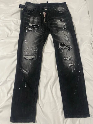 Svarta distressed jeans från Dsquared2 - Säljer ett par svarta jeans från Dsquared2 med slitna detaljer, hål och vit färgstänk. Jeansen har rak passform och dragkedja framtill med Dsquared2-logga. Perfekta för dig som gillar en edgy och trendig streetstyle.
