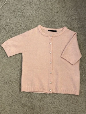 Ljusrosa kofta med pärlknappar - Supersöt ljusrosa kofta från lager 157 med korta ärmar och pärlknappar framtill. Koftan har rund halsringning och ribbade kanter. Perfekt att slänga på sig över en topp för en gullig look.