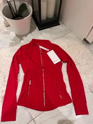 Röd zip-jacka från Lululemon - Sportig röd zip-jacka från Lululemon med figurnära passform, långa ärmar och två dragkedjeförsedda fickor. Tillverkad i ett stretchigt funktionsmaterial som passar perfekt till träning eller som ett extra lager. Snygga sömdetaljer och hög krage.