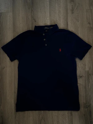 Marinblå pikétröja från Ralph Lauren - Klassisk marinblå pikétröja från Ralph Lauren med slim fit och stretch mesh-material. Tröjan har korta ärmar, krage och knappar framtill samt den ikoniska röda logotypen broderad på bröstet.