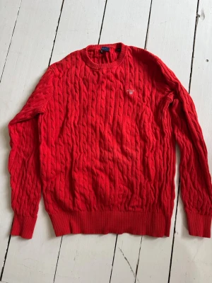 Röd kabelstickad tröja från Gant - Riktigt snygg röd kabelstickad tröja från Gant med rund halsringning och diskret logga på bröstet. Tröjan har långa ärmar och ribbade muddar vid ärmslut och nederkant. Perfekt för dig som gillar klassisk och stilren design.