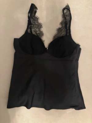 Svart spetsbody med smala axelband - Snygg svart body med tunna axelband och spetsdetaljer vid bysten. Materialet känns mjukt och följsamt, och spetsen ger en extra elegant touch. Perfekt för dig som vill ha något stilrent och trendigt i garderoben.