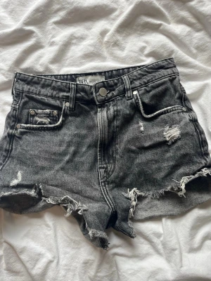 Grå denimshorts från Zara - Säljer ett par grå denimshorts från Zara med råa kanter och slitna detaljer. Shortsen har klassisk femficksdesign, knappgylf och bälteshällor. Perfekta för en avslappnad look och passar till det mesta.