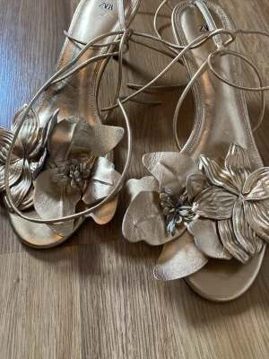 Guldiga sandaler med blommor från Zara - Säljer ett par guldiga sandaler från Zara med snörning runt ankeln och stora dekorativa blommor framtill. Sandalerna har öppen tå och är tillverkade i ett glansigt material som ger en lyxig känsla. Perfekta för att sticka ut och ge outfiten en extra touch.