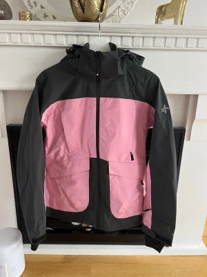 Rosa och svart skaljacka - Snygg vindjacka i rosa och svart från cross-sportwear. Jackan har huva, dragkedja framtill, två stora fickor med dragkedja och justerbara ärmslut. Tillverkad i ett lätt och tåligt material, perfekt för blåsiga dagar. Stilsäker färgkombination och sportig look. Passar perfekt för skidåkning