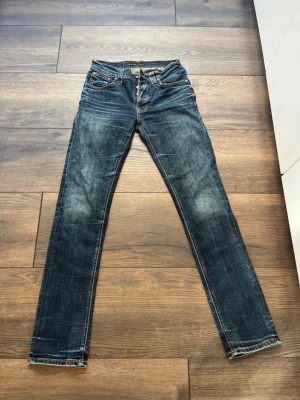 Blå slim fit jeans från Nudie Jeans - Snygga blå slim fit jeans från Nudie Jeans med klassisk femficksdesign och gul kontrastsöm. Jeansen har slitningar och en cool tvättad look, samt Nudie-logga på bakfickan. Perfekta för en avslappnad streetstil.