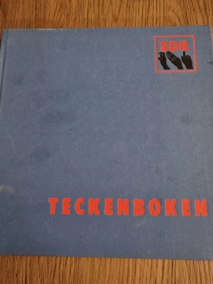 Teckenboken SDR blå hårdpärm - Teckenboken från SDR med blått hårt omslag och röd text. Boken är rektangulär och har en stilren design med SDR-logga i hörnet. Perfekt för 