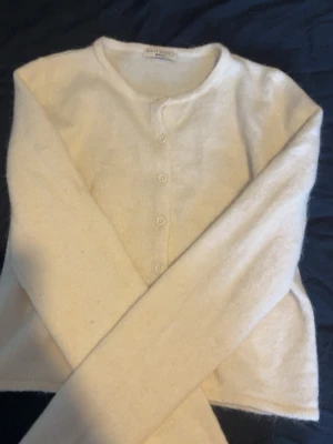 Krämvit kofta från Gina - Mysig krämvit kofta från Gina med rund halsringning och knäppning framtill. Tillverkad i mjukt, fluffigt material och har långa ärmar. Enkel och stilren design som passar perfekt till lager på lager.