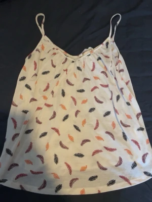 Vit linnetopp med fjädermönster - Säljer en söt vit linnetopp med tunna axelband och färgglatt fjädermönster i svart, orange och vinrött. Perfekt till sommaren och enkel att matcha med jeans eller kjol. Luftig och skön modell med avslappnad passform.
