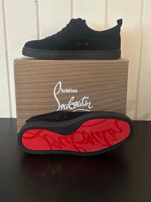 Svarta sneakers från Christian Louboutin - Stilrena svarta sneakers från Christian Louboutin med klassisk röd sula och diskreta metalldetaljer. Skorna har en låg profil och är tillverkade i mocka med snörning framtill. Perfekta för dig som gillar exklusiv och trendig streetstyle.