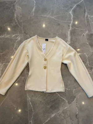 Krämvit kofta från Gina Tricot - Stilren krämvit kofta med v-ringning från Gina Tricot. Koftan har långa ärmar med guldknappar framtill. Materialet är mjukt och lätt stretchigt vilket ger en bekväm passform. 