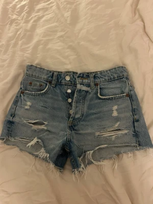 Blå håliga  jeansshorts med knappar - Säljer ett par blå denimshorts med hög midja och råa, fransiga kanter. Shortsen har slitna detaljer framtill, fyra knappar i gylfen och klassiska fem fickor. Perfekta för sommaren och ger en skön vibe.