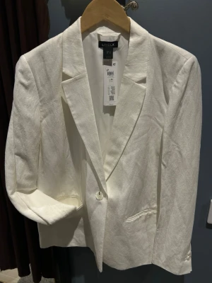 Vit linnékavaj från Lindex - Stilren vit kavaj från Lindex. Klassisk passform med spetsiga slag, enkelknäppning och två fickor framtill. Ärmarna har dekorativa knappar vid ärmslutet. Perfekt för en fräsch och trendig look. Oanvänd!