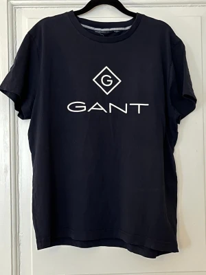 Svart GANT t-shirt med logga - Snygg svart t-shirt från GANT med stor vit logga på bröstet. Klassisk rund hals och korta ärmar. Tillverkad i mjuk bomull som känns skön mot huden. Perfekt till jeans eller shorts för en clean och enkel stil.
