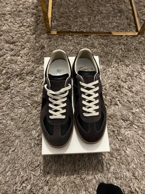 Svarta sneakers från Maison Margiela - Maison Margiela sneakers i svart mocka och läder med vita snören och klassisk gummisula. Snygg retrostil med låg profil och diskreta grå detaljer. Perfekta för dig som gillar stilrena och tidlösa sneakers. Kommer med låda och båda dustbags. Oanvända (Storlek för liten)