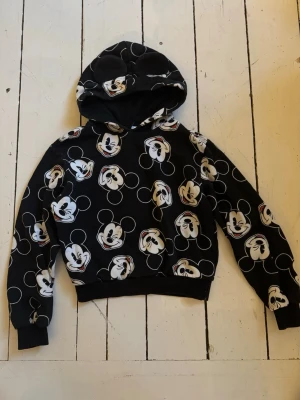 Svart hoodie med Musse Pigg-motiv Disney  - Cool svart hoodie från Disney med all-over tryck av Musse Pigg i olika uttryck. Huvan har söta öron som extra detalj och tröjan har ribbade muddar vid ärmar och midja. Perfekt för dig som älskar retro och tecknade figurer. Hoddien från Disney har storlek xs T-shirt har stl 170. Hoddien har en fläck men den syns knappt då det är så mycket motiv på den. 50 kr för både hoddie och T-shirt. 