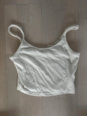 Ribbad vit crop top med spetskant - Snygg ribbad crop top/linne i vitt med tunna axelband och fin spetskant längs sidorna. Stretchigt material som sitter skönt och följer kroppen. Perfekt att matcha med jeans eller kjol för en trendig look.