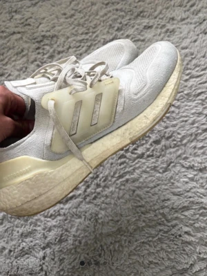Adidas vita Ultraboost sneakers - Snygga vita Adidas Ultraboost sneakers med stickad ovandel i mesh och klassiska tre ränder på sidan. Skorna har en tjock, bekväm Boost-sula och detaljer i ljusbeige plast. Perfekta för en sportig och trendig look.