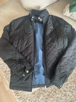 Svart quiltad jacka från Gant - Stilren svart quiltad jacka från Gant med ståkrage och tryckknappar vid halsen. Jackan har dragkedja framtill, diskret logga på bröstet och är helfodrad med blått innerfoder. Perfekt för dig som gillar klassisk och snygg stil. Pris kan diskuteras🤝