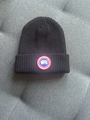 Svart ribbstickad mössa Canada Goose - Säljer en svart ribbstickad mössa från Canada Goose med klassisk uppvikt kant och det ikoniska runda Arctic Program-märket framtill. Mössan är i mjukt material och har en stilren look som passar perfekt till kalla dagar.