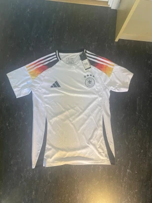 Tysklands matchtröja Adidas - Vit Tysklands matchtröja från Adidas med svarta, röda och gula detaljer på axlarna. Tröjan har korta ärmar, rund hals och tre svarta ränder på axlarna. Tysklands landslagsmärke och Adidas-logga på bröstet. Tillverkad i lätt och ventilerande material.