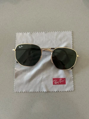 Ray-Ban guldiga solglasögon hexagon - Snygga Ray-Ban solglasögon med tunn guldfärgad metallbåge och mörka, sexkantiga linser. Klassisk och trendig modell som ger en cool vibe till din stil. Perfekta för soliga dagar och passar till de flesta outfits. Liten defekt med ena skalmen . Men inget som syns på och är lätt att fixa. 