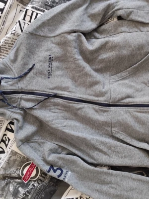 Grå hoodie från Race Marine - Snygg grå hoodie från Race Marine med dragkedja framtill och blå detaljer. Tryck på bröstet och ärmen, samt justerbar huva med snören. Perfekt för en avslappnad och sportig stil. Mjuk bomullsblandning som känns skön mot huden.