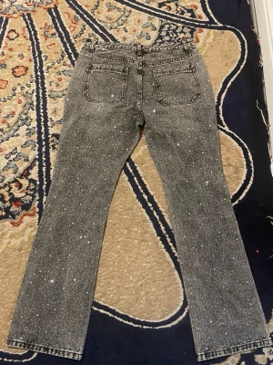 Grå jeans med strassdetaljer - Snygga grå jeans med glittrande strassdetaljer över hela tyget. Jeansen har två bakfickor och är tillverkade i en mix av bomull, polyester och viskos. Perfekta för dig som vill sticka ut med en trendig look.