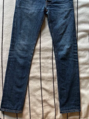 Mörkblå jeans från Nudie Jeans - Snygga mörkblå jeans från Nudie Jeans med klassisk femficksdesign och raka ben. Jeansen har subtila slitningar och kontrastsömmar samt Nudie-logga på bakfickan. Tillverkade i denim av bomull för en bekväm känsla.