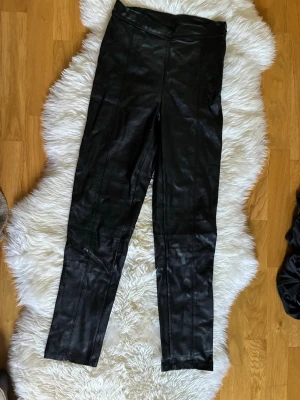 Svarta fuskskinnleggings Gina Tricot - Snygga svarta leggings i fuskskinn från Gina Tricot. De har hög midja och smal passform som sitter tight längs benen. Perfekta för att ge en edgy look till din outfit. Materialet är glansigt och ger en cool läderkänsla.